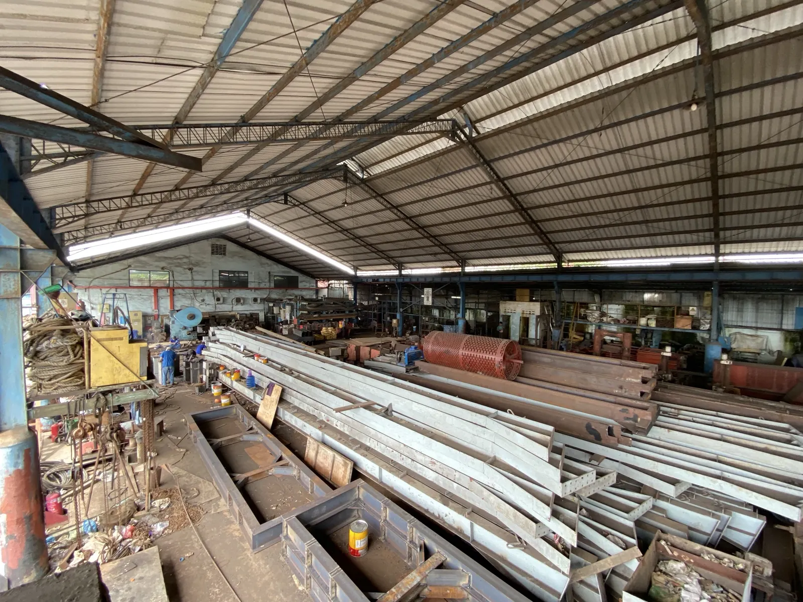 Steel beams stacked in the workshop, with a perforated red drum and machinery behind them